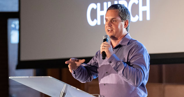 Man in purple shirt with microphone, speaking at pulpit