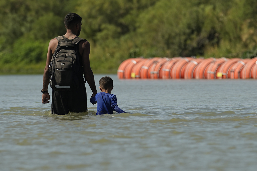 Migrants walk past large buoys being used as a floating border barrier on the Rio Grande